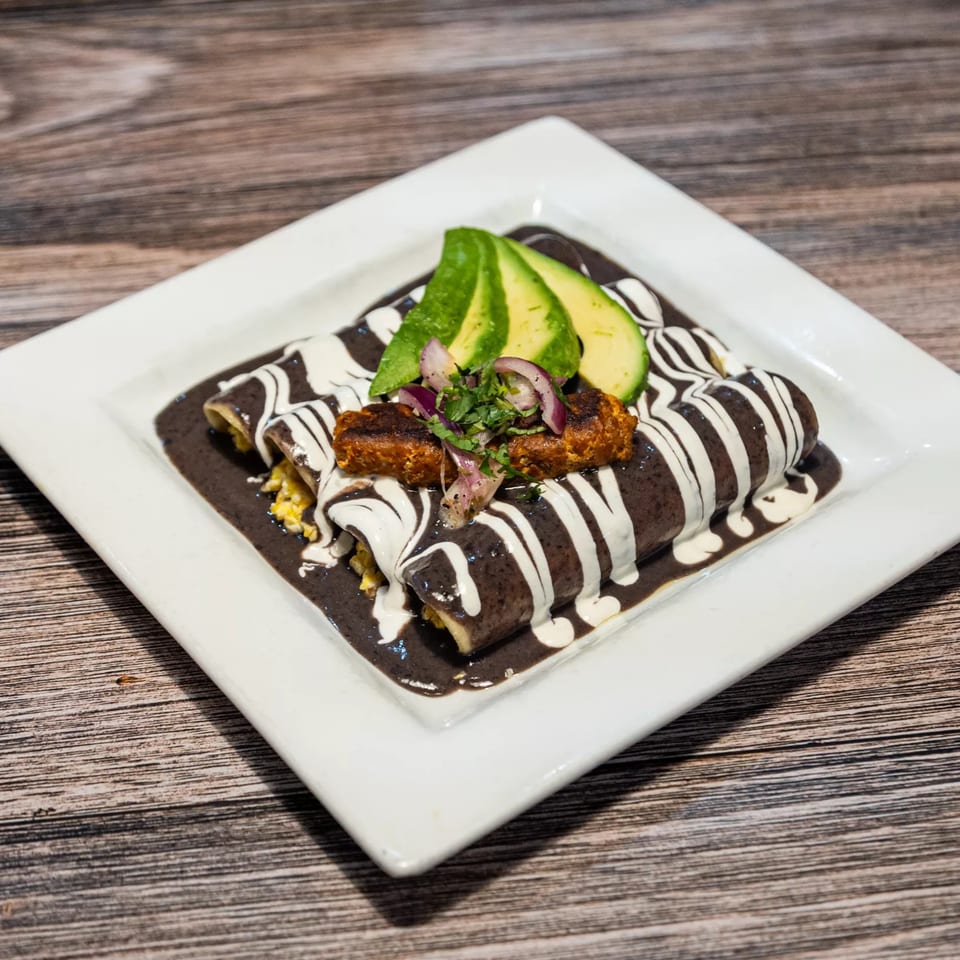 Three enchiladas covered in dark mole sauce and white crema, topped with avocado and red onion.