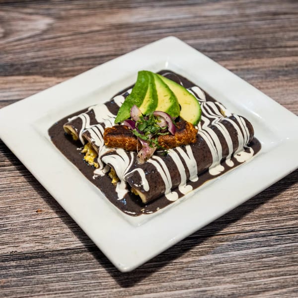 Three enchiladas covered in dark mole sauce and white cream, topped with avocado and red onion.